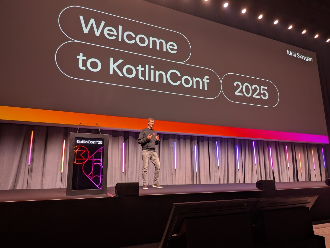 1000025514 Een spreker staat op het podium voor een groot scherm met de tekst "Welcome to KotlinConf 2025". Dit is het startschot voor een spannend conferentie-evenement gewijd aan alles wat met Kotlin te maken heeft.