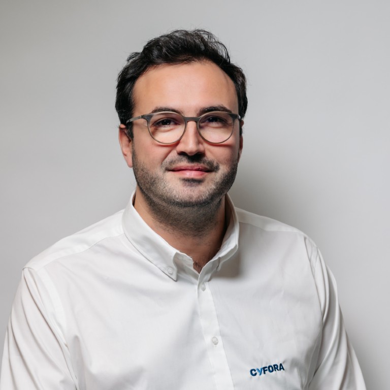 Man met kort donker haar en bril draagt een wit shirt met het Cyfora-logo tegen een lichte achtergrond.