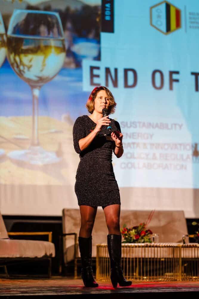 Een vrouw in een zwarte jurk en laarzen staat op het podium en houdt een microfoon vast. Ze spreekt over de datacentermarkt België voor een scherm waarop een presentatie en een afbeelding van een wijnglas te zien zijn.