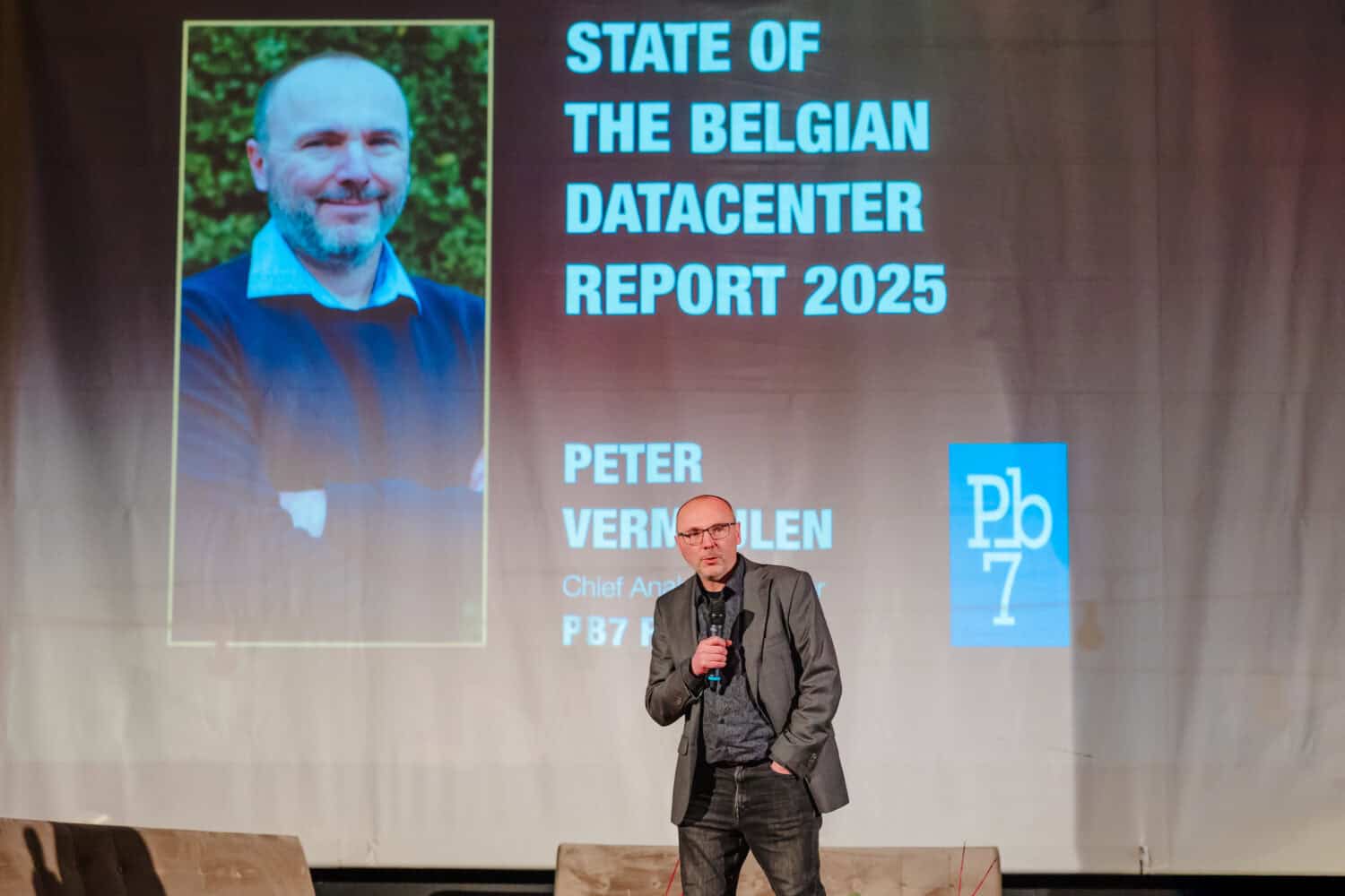 Een man staat op het podium met een microfoon voor een scherm waarop "State of the Belgian Datacenter Report 2025" te zien is, met Peter Vermuelen en inzichten in de datacentermarkt België.