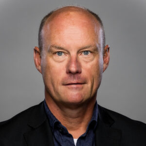 Kale man in een donker pak en blauw overhemd poseert tegen een effen grijze achtergrond, kijkt recht in de camera met een neutrale uitdrukking.