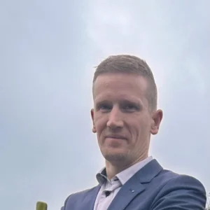 Een man in een blauw pak staat buiten met zijn armen over elkaar en kijkt naar de camera. Er is gras, een hekpaal en bomen op de achtergrond onder een bewolkte hemel.