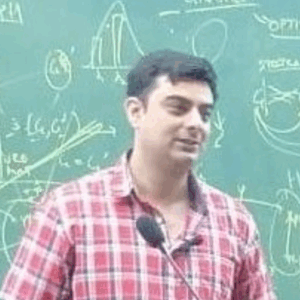 Een man in een rood geruit overhemd staat voor een krijtbord vol diagrammen en vergelijkingen en spreekt in een microfoon.