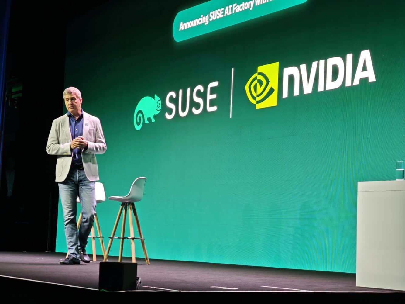 Een spreker staat op het podium naast een stoel voor een groene achtergrond met de logo's van SUSE en NVIDIA en de tekst "Announcing SUSE AI Factory with NVIDIA" (Aankondiging SUSE AI Factory met NVIDIA) tijdens SUSECON, waarbij de nadruk wordt gelegd op vooruitgang in AI en digitale soevereiniteit.