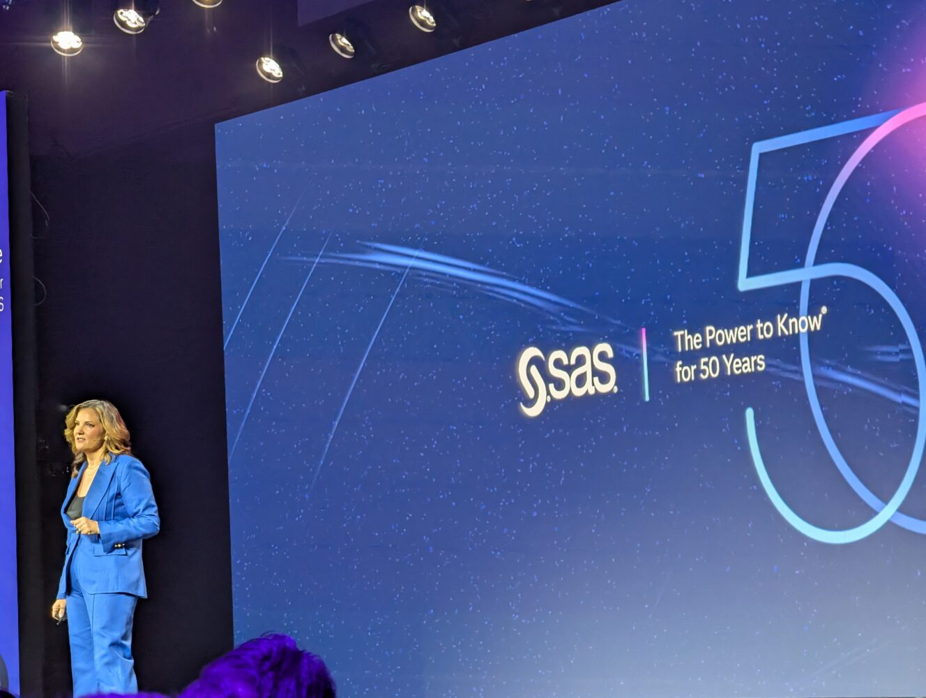 Een vrouw in een blauw pak staat op het podium naast een groot scherm met het SAS-logo en de tekst "The Power to Know for 50 Years," waarmee de nalatenschap van SAS op het gebied van analytics wordt gevierd.