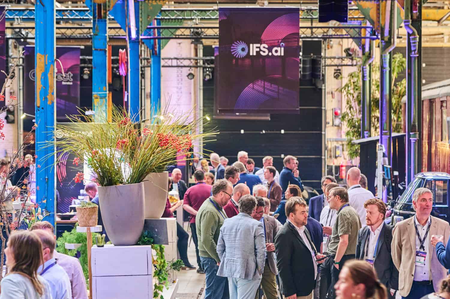 Een menigte mensen in zakelijke kleding verzamelt en praat op een indoor evenement met IFS.ai en IFS Nexus Black banners erboven.