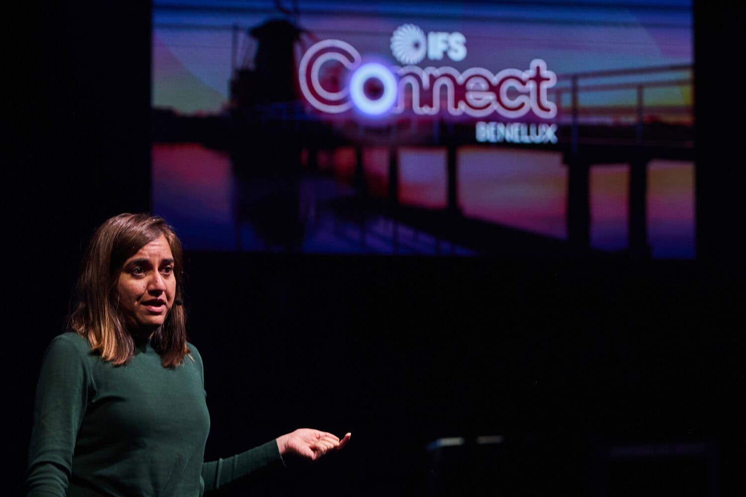 Een vrouw in een groene trui spreekt op het podium voor een scherm met het logo van IFS Connect Benelux en het merk IFS Nexus Black.