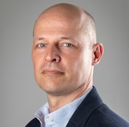 Kale man in een donker colbert en lichtblauw overhemd poseert tegen een effen grijze achtergrond.