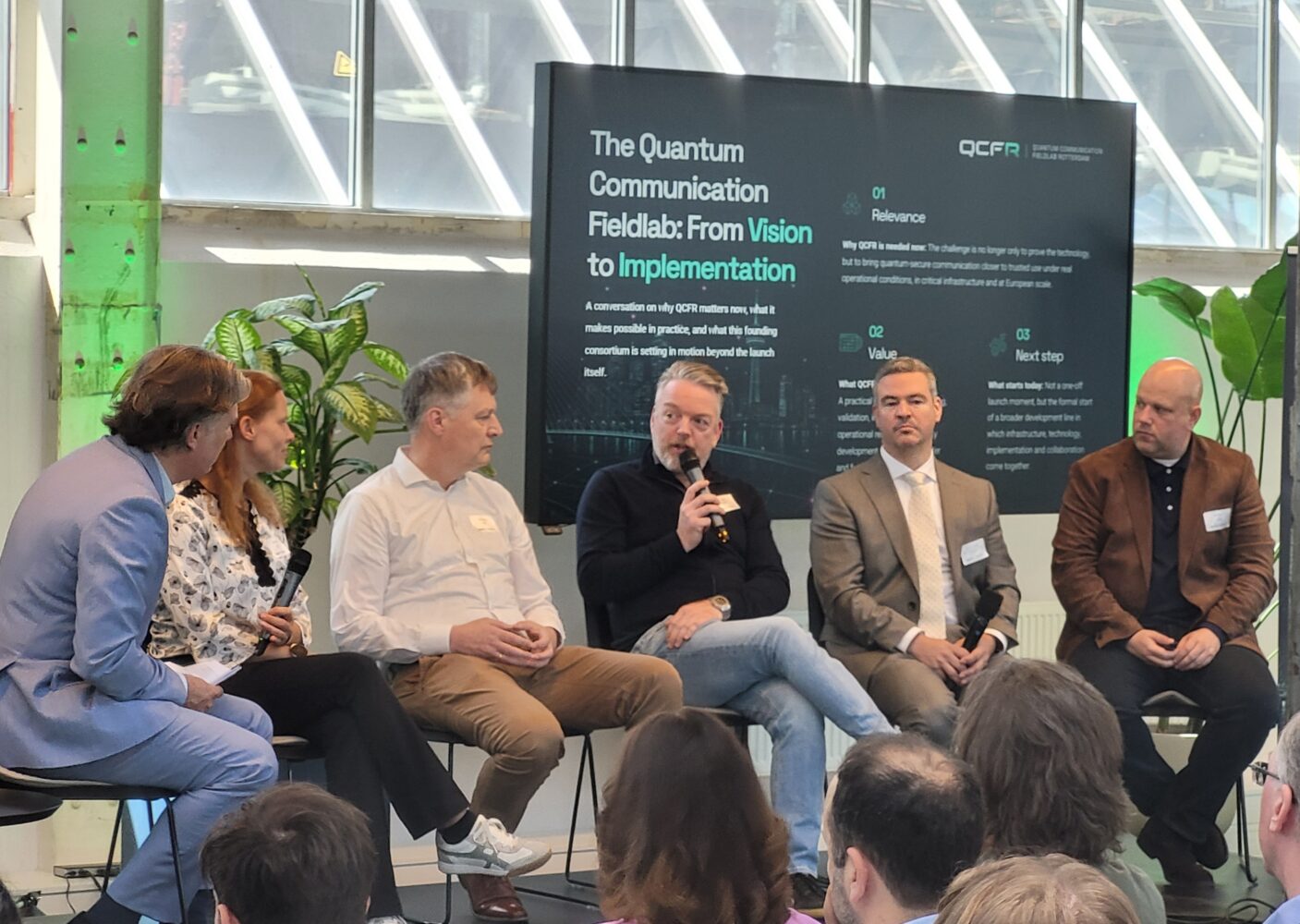 Marc Hulzebos panel QCFR event R’dam 1 Zes mensen zitten op het podium voor een publiek, terwijl één persoon in een microfoon spreekt. Op een groot bord achter hen staan de woorden ‘kwantumveilig veldlaboratorium Rotterdam’ en ‘implementatie van kwantumcommunicatie’.